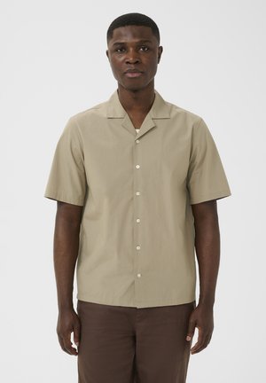 Noor mees seisab näoga otse vaataja poole, kandes lühikeste varrukatega beežikat nööpidega särki ja tumedat pruunikat püksipaaritust ühetaustalise heleda tausta ees.