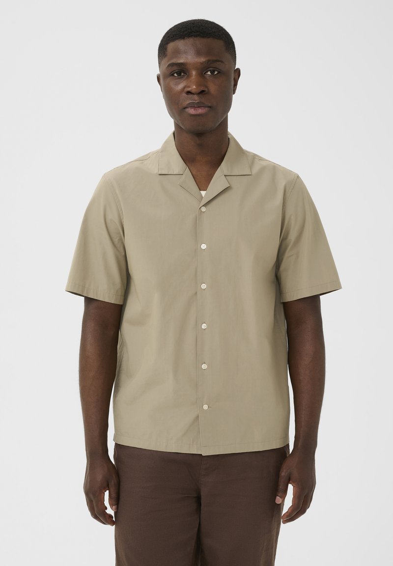 Un jeune homme se tient face à la caméra, portant une chemise beige à manches courtes et un pantalon marron foncé, sur un fond clair uni.