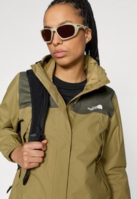 Αδιάβροχο μπουφάν ελιάς με μαύρες λεπτομέρειες στους ώμους και λογότυπο The North Face, συνδυασμένο με γυαλιά ηλίου σε ανοιχτό ταμπά χρώμα και μαύρο σακίδιο πλάτης.