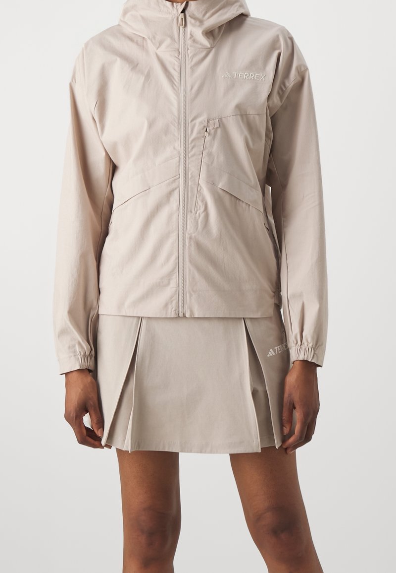 Veste beige légère avec capuche, fermeture éclair à l'avant et poches latérales, associée à une jupe plissée assortie avec des accents de logo.