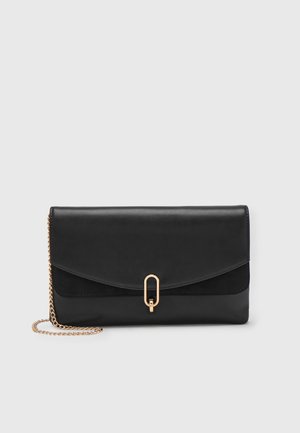 Pochette en cuir noir avec bandoulière chaîne dorée et fermoir ovale doré, sur fond uni gris clair.