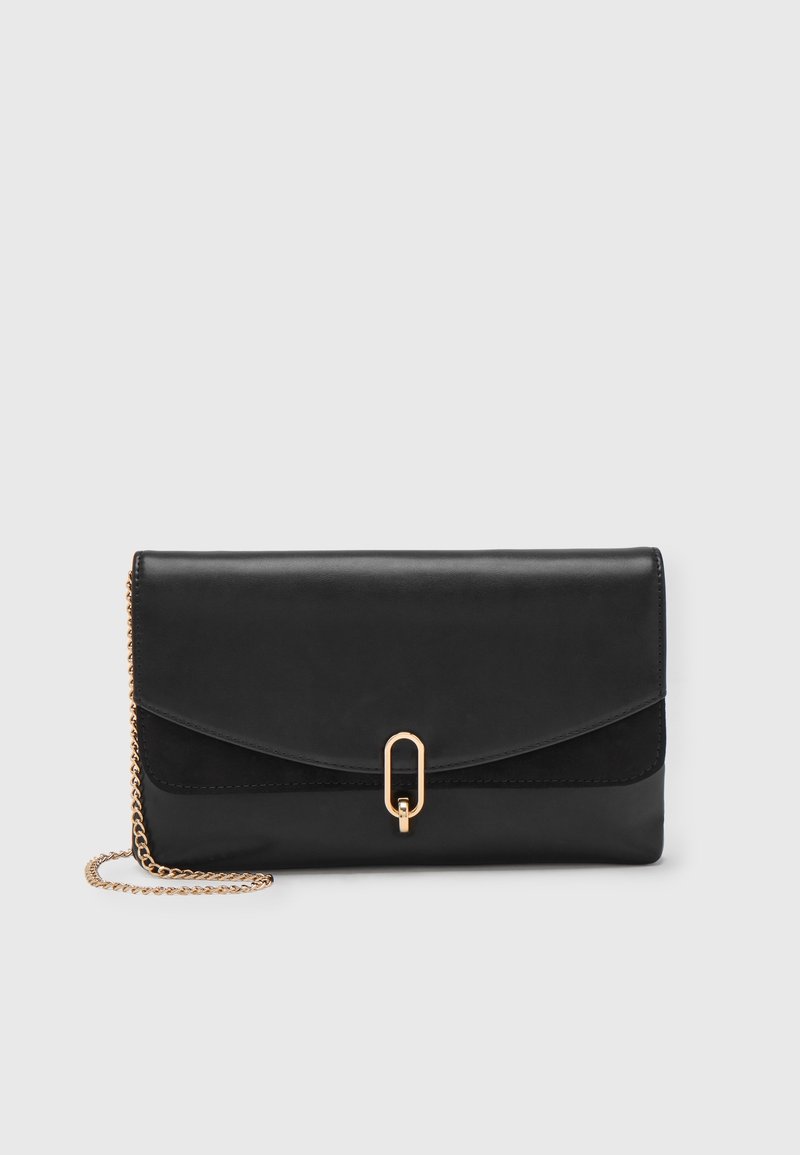 Pochette en cuir noir avec bandoulière chaîne dorée et fermoir ovale doré, sur fond uni gris clair.