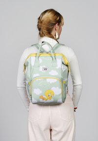 Mochila verde con acentos amarillos, que presenta un patrón de pájaros de dibujos animados y nubes. Hecha de un material suave, con dos asas superiores y bolsillos con cremallera.