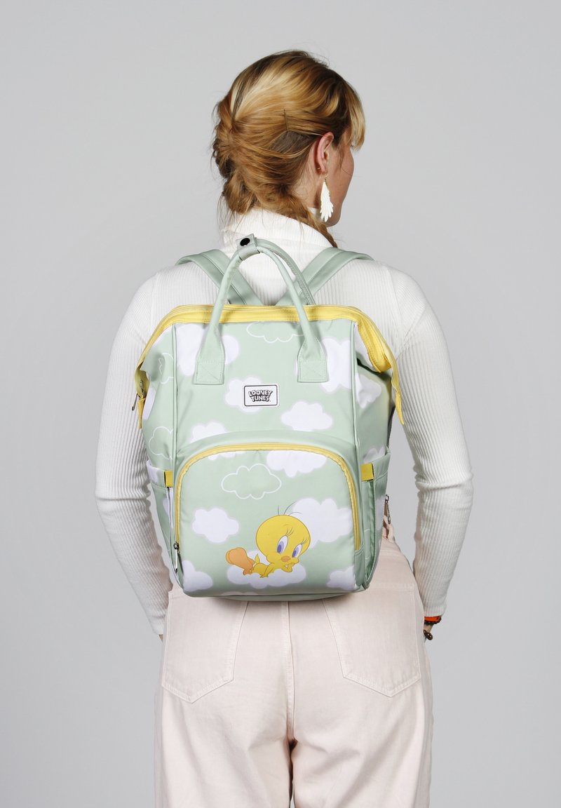 Mochila verde con acentos amarillos, que presenta un patrón de pájaros de dibujos animados y nubes. Hecha de un material suave, con dos asas superiores y bolsillos con cremallera.