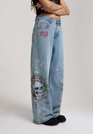 Person iført lyseblå vidde jeans med kranie- og blomsterprint og rød broderet tekst, parret med sorte sko, stående med armene over kors.