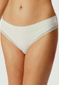 Žena v bílém bavlněném spodním prádle ve stylu bikin s krajkovým lemem na pase a na nohavičkách, stojící na jednobarevném pozadí.