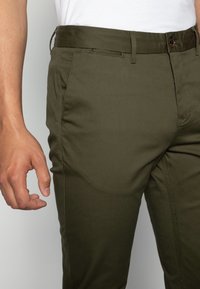 Pantaloni verde oliva con una texture liscia, fit affusolato e tasche laterali. Presentano un bottone marrone e passanti per cintura in vita.