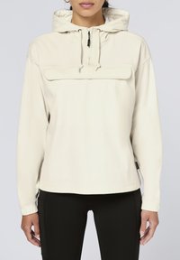 Light beige hooded pullover made from ribbed fabric, featuring a front pouch pocket and elastic cuffs, paired with black leggings.