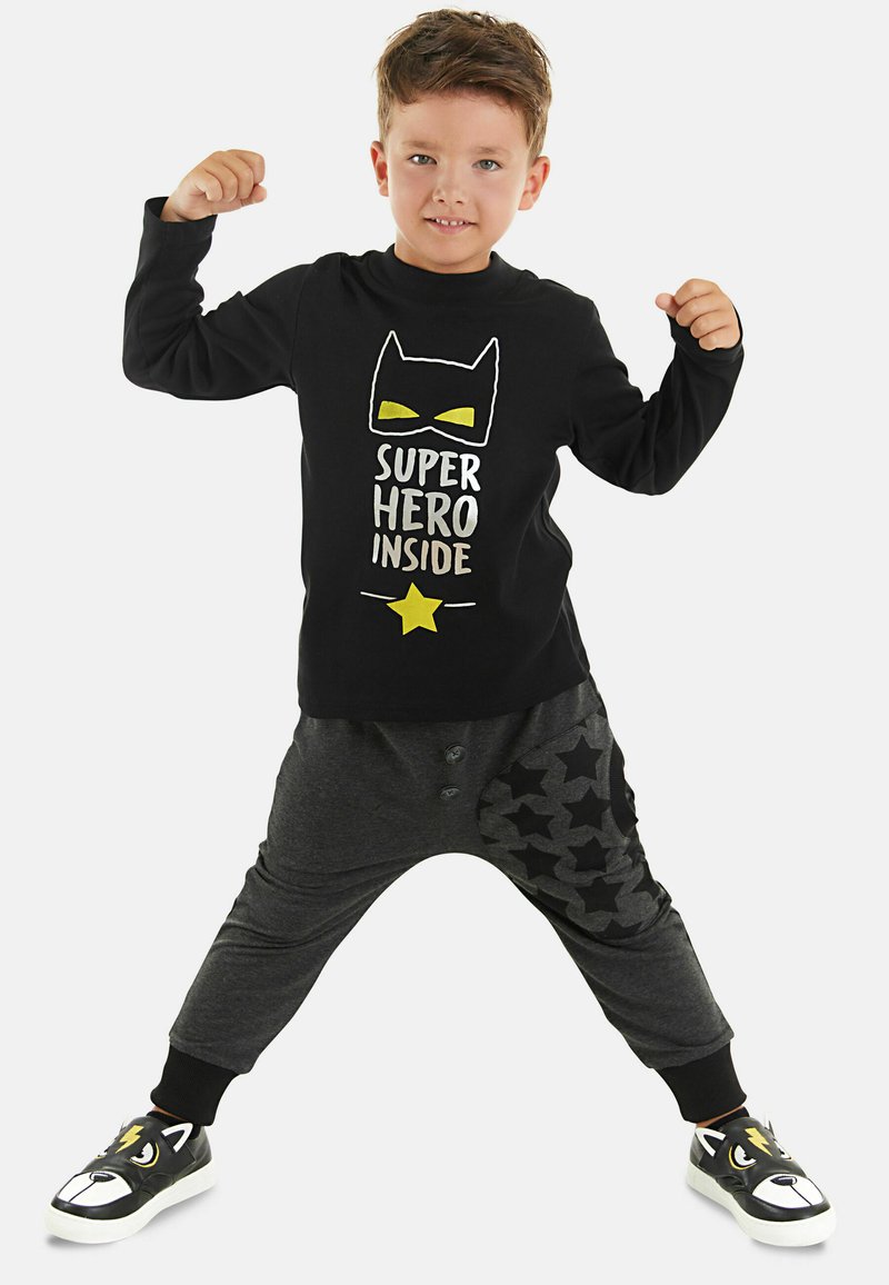 Maglietta nera a maniche lunghe con maschera da supereroe gialla e testo; pantaloni jogger grigi con motivo a stelle; sneakers nere con design di faccia di animale.