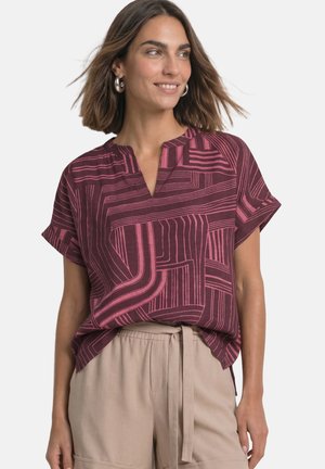 Femme aux cheveux jusqu'aux épaules portant un haut bordeaux à manches courtes avec des lignes géométriques roses et un pantalon beige à cordon, souriante et regardant vers la droite.
