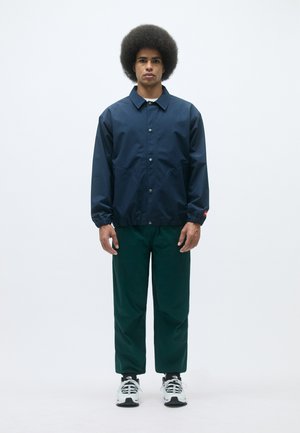 Casaco impermeável azul-marinho com botões de pressão, punhos elásticos e corte casual, combinado com calças verdes e ténis preto e branco.