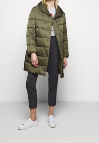 Puffet olivengrøn frakke med stor hætte, mat finish og lodrette syninger. Kombineret med grå bukser og hvide sneakers.