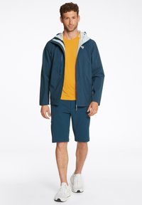 Chaqueta navy resistente al agua con capucha gris claro, combinada con una camiseta naranja brillante y pantalones cortos navy oscuros. Zapatillas blancas completan el look.