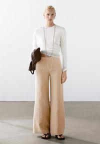 Voorzien van ruime beige linnen broek met verticale strepen, gecombineerd met een witte lange mouwen top en zwarte sandalen. Accessoires omvatten een gestructureerde bruine tas.