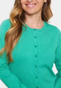 Roheline soonikuga kardigan, millel on ümmargune kaelus, pikad varrukad ja keskel nööbistamine. Sujuv tekstuur ja kohandatud disain.