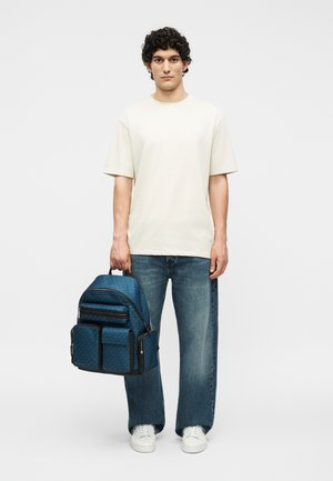 Giovane uomo con una maglietta bianca e jeans che tiene uno zaino blu con più tasche frontali e cerniere nere.