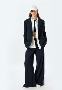Marineblauwe oversized blazer met twee knopen, witte blouse, zwarte crop top, wijde marineblauwe broek en een lichtgrijze pet.