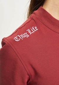 Burgunderfarvet t-shirt med ribbet krave, der har hvid broderet "Thug Life" tekst på venstre skulder, glat tekstur og tætsiddende design.