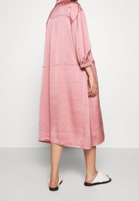 Long, loose-fitting dress in soft pink fabric with balloon sleeves and a curved hem, paired with white slip-on sandals.