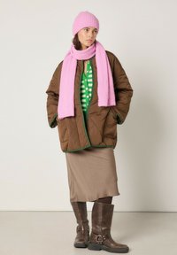 Bruine donsjas met groene bies, roze sjaal en hoed, groene gestreepte trui, beige rok, en bruine kniehoge laarzen met gespen.