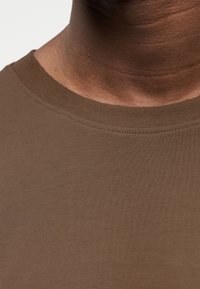 Καφέ μπλούζα με στρογγυλή λαιμόκοψη, με λεία υφή και εφαρμοστή σχεδίαση, που δείχνει λεπτομέρειες ραφής κατά μήκος της λαιμόκοψης και της επιφάνειας του υφάσματος.