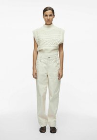 Top sin mangas de punto en un tono crema claro con patrones de trenzado, combinado con pantalones de pierna ancha en crema de cintura alta. Sandalias negras de tiras completan el look.