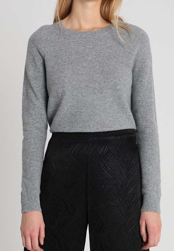 Femme portant un pull gris ajusté à manches longues, rentré dans un pantalon noir taille haute avec un motif texturé subtil.