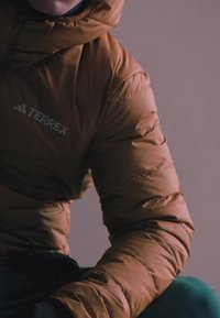 Henkilö, joka istuu yksinkertaisen vaalean taustan edessä ja pukeutuneena ruskeaan Terrex-toppatakkiin, jossa on huppu.