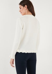Weißer, strukturierter Pullover mit Rundhalsausschnitt, gerafftem Saum und langen Ärmeln, der eine figurbetonte Silhouette und dezente Details aufweist.