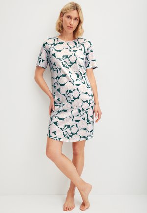 Donna in piedi a piedi nudi contro un muro bianco, che indossa un vestito a maniche corte con grandi motivi floreali bianchi su uno sfondo verde scuro.