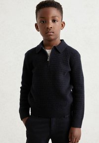 Mornarsko plavi pleteni pulover s ovratnikom na patentni zatvarač, s teksturiranim uzorkom, dugim rukavima i rebrastim manžetama. Uparen s tamnim hlačama.