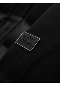 Tejido negro con una placa metálica rectangular en negro y plata etiquetada "OMBRE EXIMIO" adjunta a la superficie.