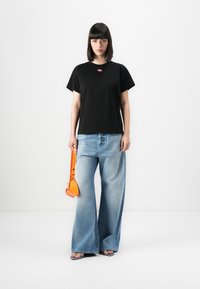 Woman with black bob haircut wearing black t-shirt, wide-leg faded jeans, holding an orange handbag, standing on white background.