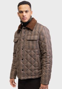 Mand iført brun ternet quiltet jakke med corduroy krave og sort turtleneck, stående mod en ensfarvet lys baggrund.