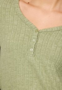 Lys grøn ribbet top med V-udskæring, tre knapper i midten og en blød tekstur. Stoffet ser let og åndbart ud.