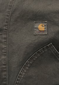 Σκούρο γκρι ύφασμα με λεπτομερείς ραφές δείχνει ένα μικρό τετράγωνο λογότυπο Carhartt πάνω από την καμπυλωτή άκρη της τσέπης.