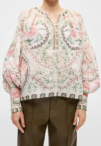 Blouse ample à manches longues avec motifs floraux et feuillages en rose, vert et marron, portée avec un pantalon olive foncé.
