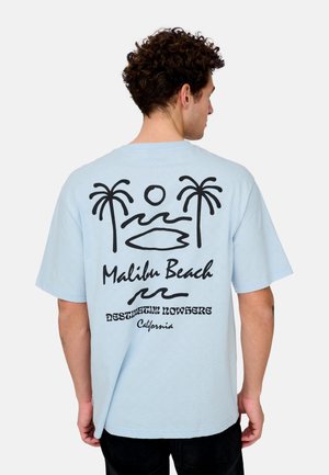 Hombre con cabello corto y rizado que lleva una camiseta azul claro con palmeras negras, sol, olas, tabla de surf y el texto "Malibu Beach, Destination Nowhere, California."