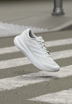 Scarpa da corsa bianca con tomaia in mesh e tre strisce, sospesa in aria sopra un attraversamento pedonale in una strada urbana.