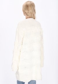 Vit stickad cardigan med strukturerade, vågiga fransdetaljer. Långa ärmar, öppen front och en avslappnad passform. Mjuk och lättviktsmaterial.