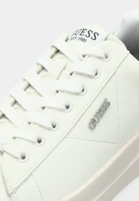 Weiße synthetische Sneaker mit glatter Textur, ausgestattet mit sechs weißen Schnürsenkeln, einem silbernen "GUESS"-Logo und einem dreieckigen Akzent an der Seite.