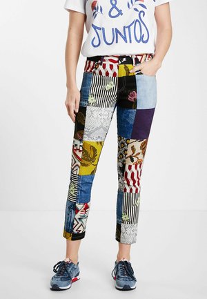 Kvinde iført patchworkmønstrede bukser med forskellige farver og designs, hvid t-shirt med grafik og blå sneakers, stående med den ene hånd i lommen.