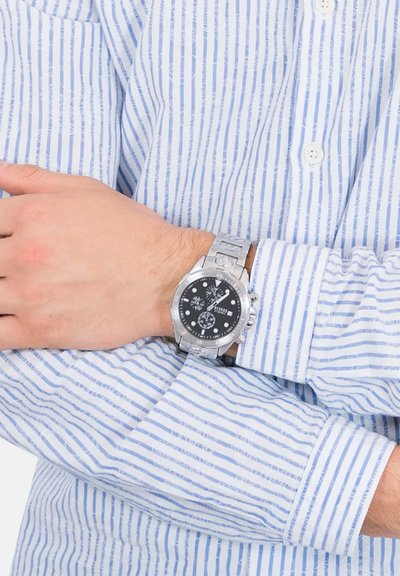 Orologio in acciaio inossidabile argentato con quadrante nero, indicatori delle ore bianchi a contrasto e bracciale a maglie, indossato su un polso coperto da una camicia a righe.