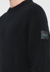 Pulover negru din tricot, cu guler rotund ribetat și o etichetă țesută pe mâneca stângă, prezentând un detaliu cu logo dreptunghiular.