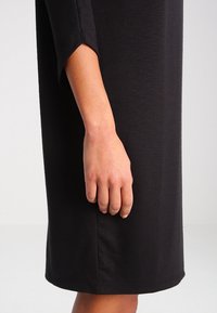 Robe noire mi-longue en tissu doux, avec des manches trois-quarts à coupe ample. Détails de coutures visibles sur le côté.