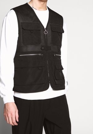 Sort teknisk vest med flere forlommer og sølvfarvede lynlåse, båret over en hvid langærmet skjorte og sorte bukser.