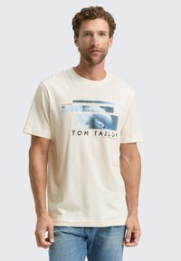 Hombre con cabello rizado que lleva una camiseta gráfica beige con formas abstractas azules y texto, combinada con jeans azules, de pie contra un fondo blanco.