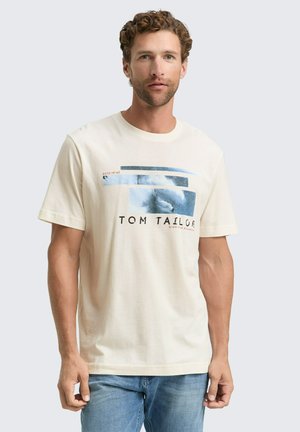 Mann mit lockigem Haar, der ein beiges Grafik-T-Shirt mit blauen abstrakten Formen und Text trägt, kombiniert mit blauen Jeans, steht vor einem weißen Hintergrund.