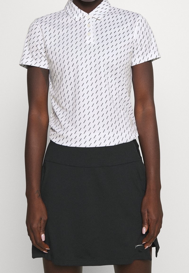 Personne portant un polo blanc à manches courtes avec des lignes diagonales noires et une jupe noire avec un petit logo près de l'ourlet.