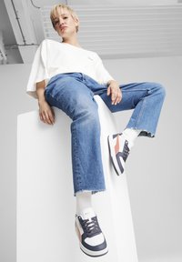 Sneaker aus weißem Leder mit navyblauen Akzenten und korallenfarbigen Details. Das Modell trägt ein weißes Oversize-Shirt und ausgewaschene blaue Jeans.